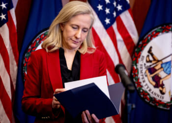 Abigail Spanberger in a red jacket looks down at a file. Behind her, two American flags and a Virginia state flag.