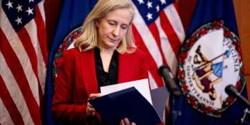 Abigail Spanberger in a red jacket looks down at a file. Behind her, two American flags and a Virginia state flag.