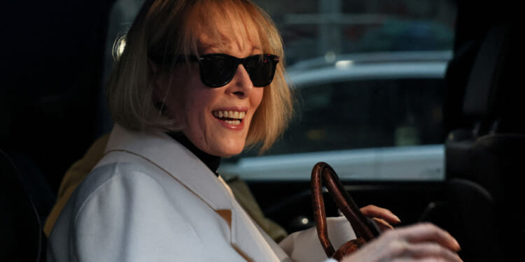 Close-up of E. Jean Carroll in sunglasses and white coat smiling while seated in car.