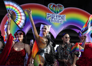 Colorfully-dressed individuals celebrate in front of a rainbow backdrop reading "Love PRIDE 2026)