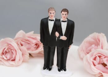 A wedding cake with a decoration showing two grooms on top of it.