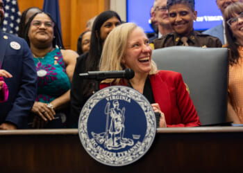 A laughing Abigail Spanberger sits at a desk surrounded by smiling, laughing people.