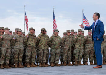 Secretary of War Pete Hegseth points as he's standing with and addressing National Guard troops with numerous American flags behind them.