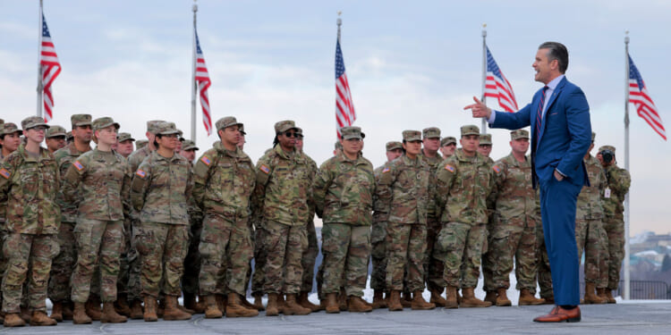 Secretary of War Pete Hegseth points as he's standing with and addressing National Guard troops with numerous American flags behind them.