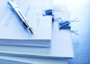 Stacks of paperwork sit on a table with a ballpoint pen topping the stack.