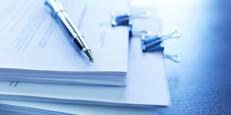 Stacks of paperwork sit on a table with a ballpoint pen topping the stack.