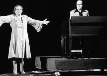 Black-and-white photo of singer Kate Smith on stage with her arms spread wide. To her left a man on keyboards.