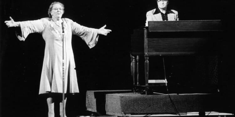 Black-and-white photo of singer Kate Smith on stage with her arms spread wide. To her left a man on keyboards.