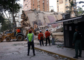 Several workers stand in front of the rubble of a collapsed building as a bulldozer works with the rubble.