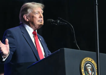 Side view of Donald Trump at podium against a dark background with his right arm gesturing to the side.