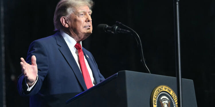 Side view of Donald Trump at podium against a dark background with his right arm gesturing to the side.