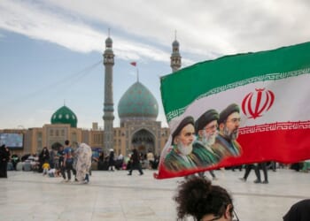 People hold a flag with Iran's late and new supreme leaders, Ayatollah Ruhollah Khomeini, Ayatollah Ali Khamenei, and Ayatollah Mojtaba Khamenei, during Eid al-Adha at the entrance of Jamkaran Mosque on May 26, 2026, in Qom, Iran.