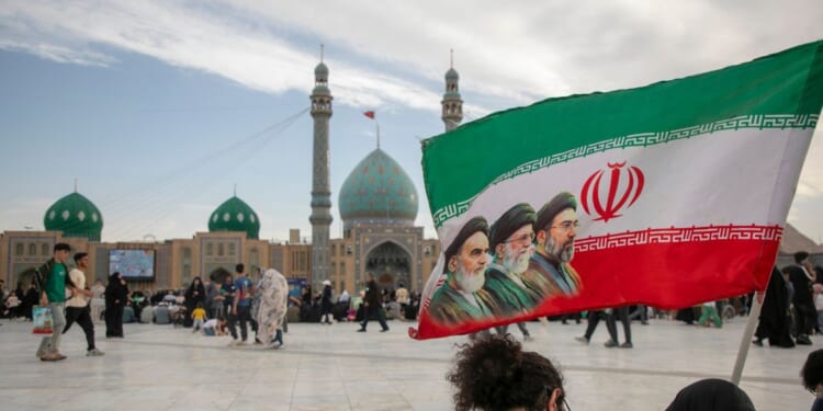 People hold a flag with Iran's late and new supreme leaders, Ayatollah Ruhollah Khomeini, Ayatollah Ali Khamenei, and Ayatollah Mojtaba Khamenei, during Eid al-Adha at the entrance of Jamkaran Mosque on May 26, 2026, in Qom, Iran.