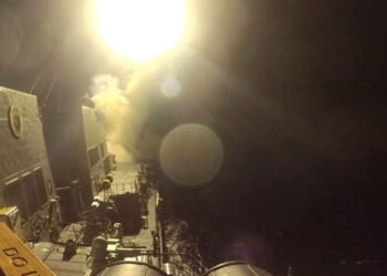 A missile is launched from a naval ship at night, creating a bright burst of light and smoke against the dark sky and ocean. The ships deck and equipment are partially visible under the intense glow.