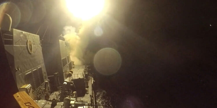 A missile is launched from a naval ship at night, creating a bright burst of light and smoke against the dark sky and ocean. The ships deck and equipment are partially visible under the intense glow.