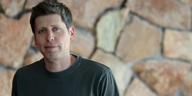 A man with short brown hair wearing a dark green shirt stands in front of a wall made of large, irregular stone blocks.