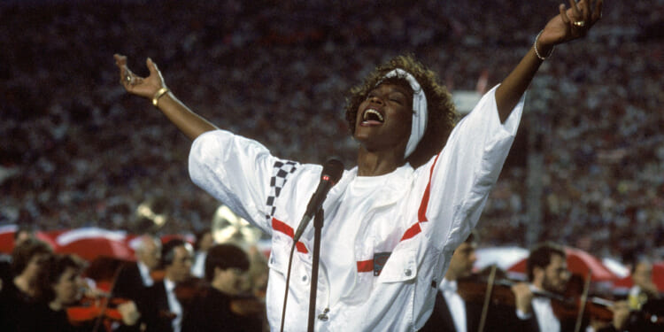 Whitney Houston in a white, with blue and red trim pullover, has her arms outstretched as she concludes singing "The Star-Spangled Banner" during Super Bowl XXV