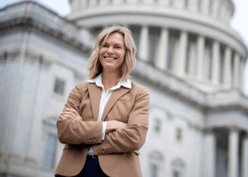 A woman in a tan blazer stands confidently with arms crossed in front of a government building with columns and a dome, smiling at the camera.