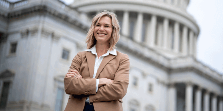 A woman in a tan blazer stands confidently with arms crossed in front of a government building with columns and a dome, smiling at the camera.