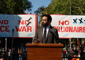 Zohran Mamdani stands at a podium speaking at a rally, with banners behind him reading NO ICE, NO WAR, and NO BILLIONAIRES.