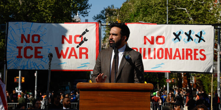Zohran Mamdani stands at a podium speaking at a rally, with banners behind him reading NO ICE, NO WAR, and NO BILLIONAIRES.
