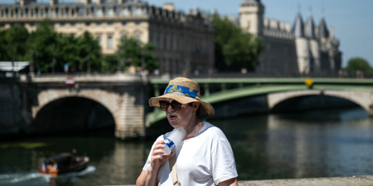 A woman with a hat cools off with a cold bottle of water on the Seine riverside with the Paris commercial court (L) and the Conciergerie buildings and the Pont Notre Dame in the background as France experiences a heat wave in Paris, France, on June 21, 2026.
