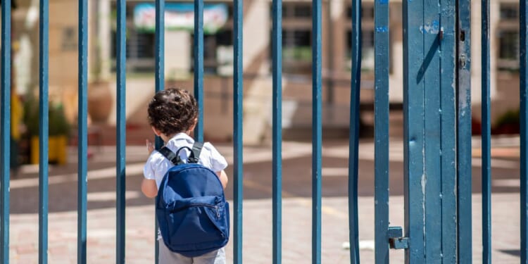A child with a school backpack look on schoolyard grounds toward an open entrance door.