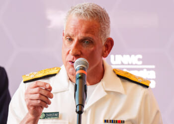 A person in a white military uniform with gold shoulder boards speaks into a microphone at a podium, with a blurred purple background and visible name tag reading “Christine.”.