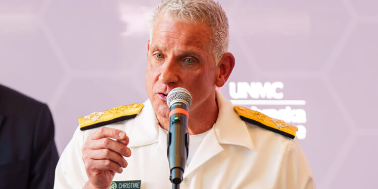 A person in a white military uniform with gold shoulder boards speaks into a microphone at a podium, with a blurred purple background and visible name tag reading “Christine.”.
