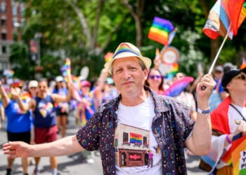Brad Lander smiling and wearing a rainbow hat and patterned shirt stands at a Pride parade, surrounded by people waving rainbow flags and celebrating. Trees and buildings are visible in the background.