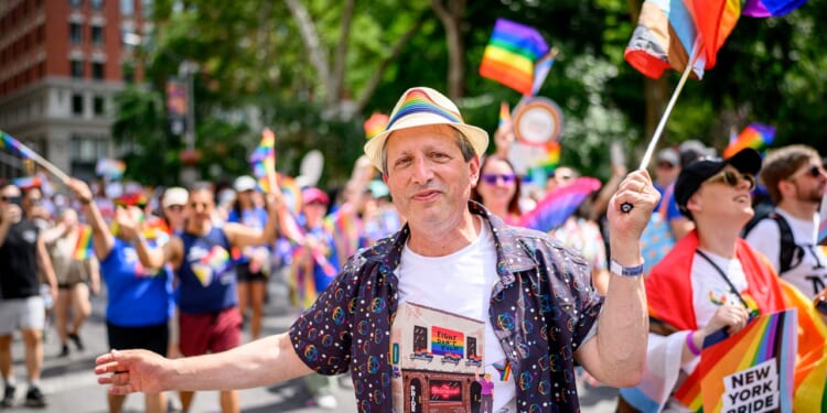 Brad Lander smiling and wearing a rainbow hat and patterned shirt stands at a Pride parade, surrounded by people waving rainbow flags and celebrating. Trees and buildings are visible in the background.