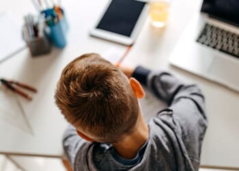 A boy does school work on a desk next to a computer, tablet and various schools supplies.