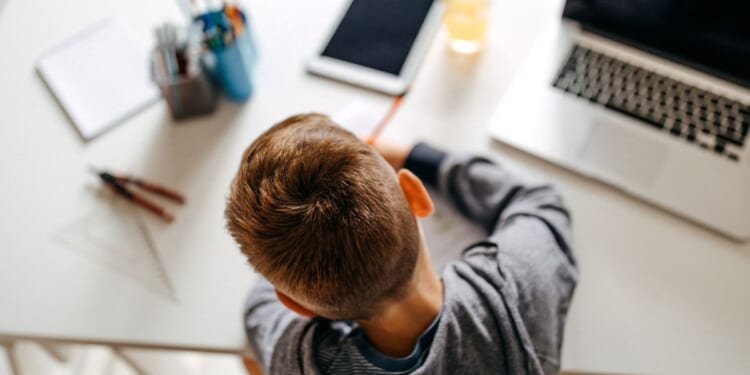 A boy does school work on a desk next to a computer, tablet and various schools supplies.