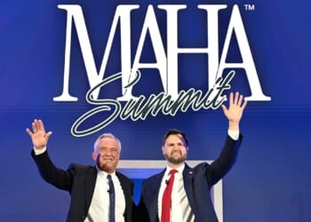 Two men in suits stand side by side, smiling and waving, in front of a blue backdrop with large white text that says “MAHA Summit.”.