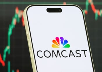 The Comcast logo is displayed on a phone while a stock market depiction is pictured behind.