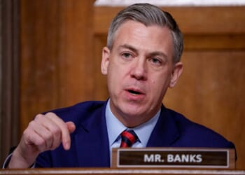 A man in a navy suit and striped tie speaks, gesturing with his hand, seated behind a nameplate that reads Mr. Banks in a wood-paneled room.