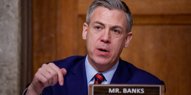 A man in a navy suit and striped tie speaks, gesturing with his hand, seated behind a nameplate that reads Mr. Banks in a wood-paneled room.