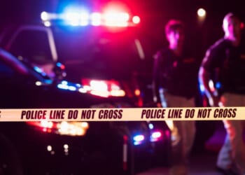 Two police officers standing in front of patrol cars, behind police tape, wearing bulletproof vests and duty belts.