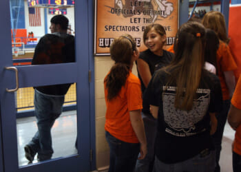 A group of girls in orange and black shirts stand and talk near a gym entrance.