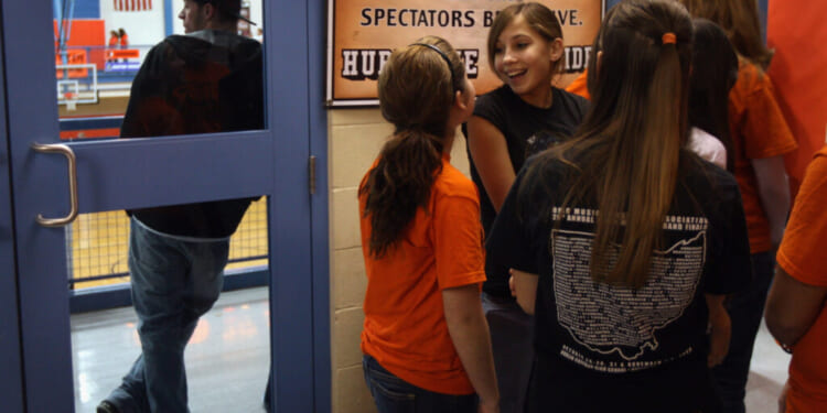A group of girls in orange and black shirts stand and talk near a gym entrance.