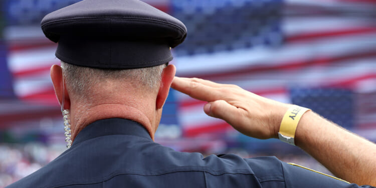 A police officer with a security wristband salutes in front of a large, blurred American flag backdrop, proudly honoring America.