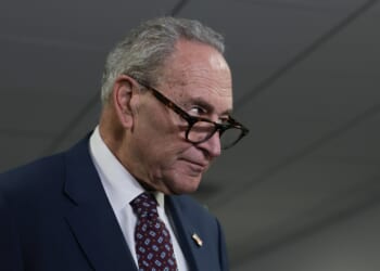 Chuck Schumer in a suit and patterned tie looks over the top of his glasses with a serious expression, standing indoors under fluorescent lights.