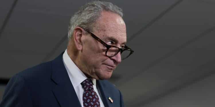 Chuck Schumer in a suit and patterned tie looks over the top of his glasses with a serious expression, standing indoors under fluorescent lights.