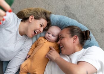A same-sex couple of women lay on the ground with a baby as it stares up at a phone.