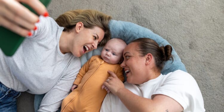 A same-sex couple of women lay on the ground with a baby as it stares up at a phone.