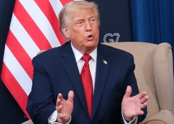 Trump in a dark suit and red tie sits in a beige chair, gesturing with his hands. Behind him are a partial U.S. flag and banners with G7 France 2019 and the U.S. State Department seal.