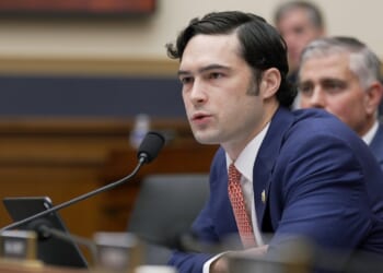 A man in a navy suit speaks into a microphone at a desk in a formal meeting room, with another man seated behind him and blurred background figures.