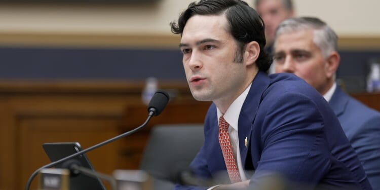 A man in a navy suit speaks into a microphone at a desk in a formal meeting room, with another man seated behind him and blurred background figures.