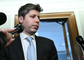 OpenAI CEO Sam Altman is surrounded by microphones, standing in front of an office doorway, appearing to speak or prepare to speak to reporters.