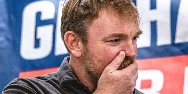 A man with light brown hair and a beard covers his mouth with his hand, appearing thoughtful or concerned. A blue banner with large white letters is blurred in the background.